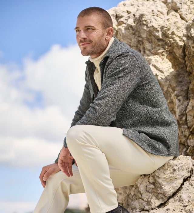 Ein Mann sitzt auf einem Felsen und trägt eine Hemd Jacke in Schwarz, darunter einen Rollkragen Pullover mit Zopfmuster in Offwhite und dazu eine helle Hose.