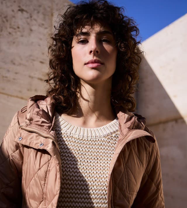 Eine Frau steht vor einer Natursteinwand und trägt eine Stepp Kurzjacke in der Farbe Praline, darunter einen passenden Pullover mit dezentem Streifen.