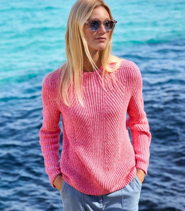 Eine blonde Frau mit Sonnenbrille steht am Wasser und trägt einen Pullover in Rosa, dazu eine Jeans.