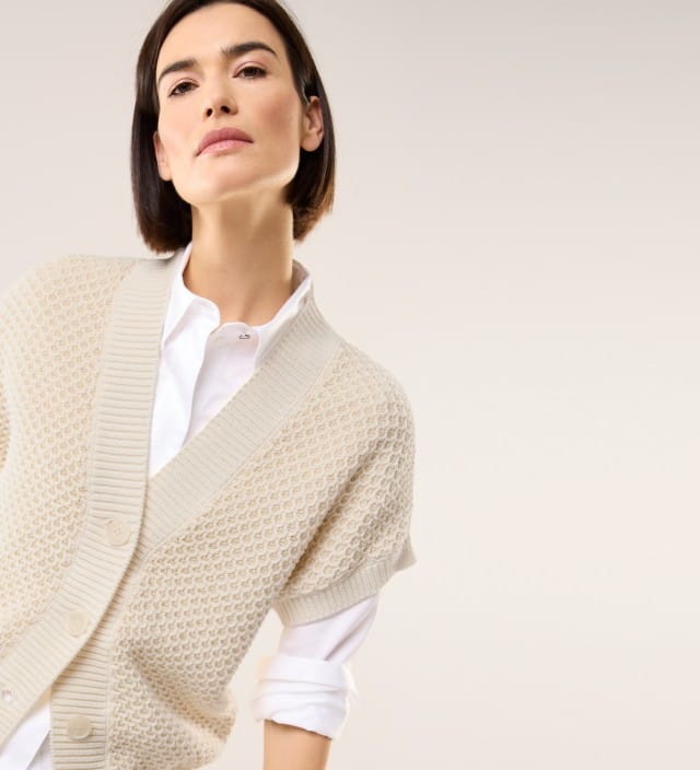 Eine Frau trägt eine weiße Bluse, darüber ein kurzärmligen Strick-Cardigan in Beige. 