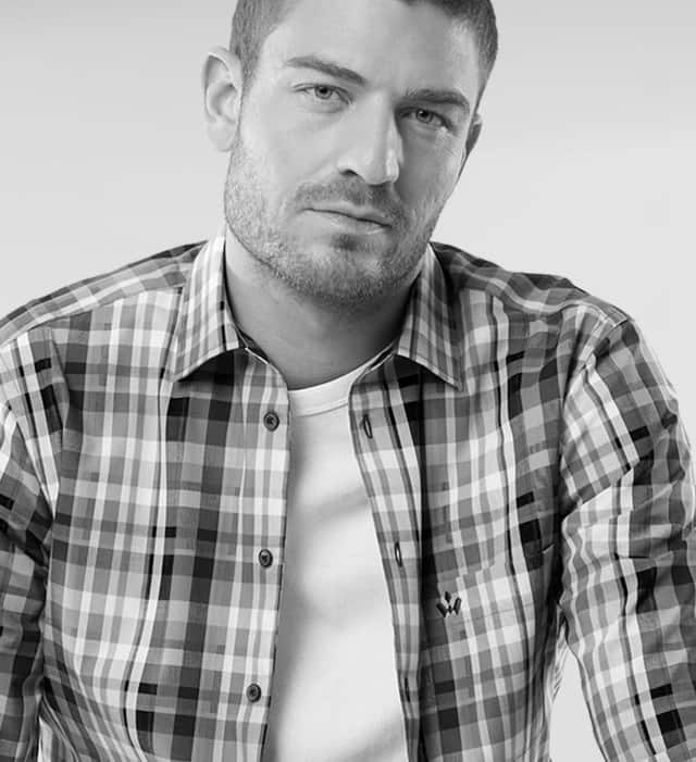 Ein Mann trägt ein offenes kariertes Hemd, darunter ein weißes Shirt.