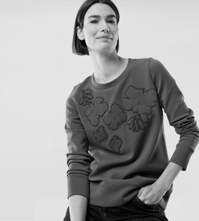 Eine Frau trägt ein Sweatshirt mit floralen Applikationen und eine Jeans. Ihr Stil wirkt modern, bequem und zugleich feminin.