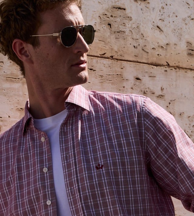 Ein Mann mit Sonnenbrille steht vor einer Natursteinwand und trägt ein rotes kariertes Hemd, darunter ein weißes Shirt. Ein Mann mit Sonnenbrille steht vor einer Natursteinwand und trägt ein rotes kariertes Hemd, darunter ein weißes Shirt.