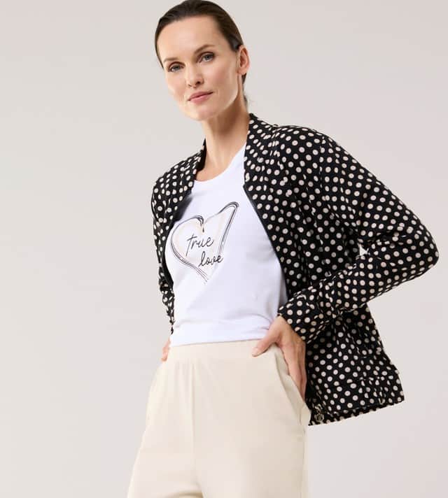 Eine Frau trägt einen gepunkteten Blouson, darunter ein weißes T-Shirt und dazu eine Hose in Beige.