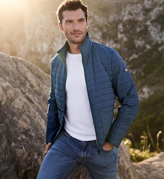 Ein Mann steht in einer Felslandschaft und trägt eine blaue Steppjacke, darunter ein weißes Shirt und dazu eine Jeans. 