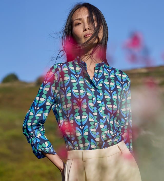 Eine Frau steht auf einer Frühlingswiese und trägt eine gemusterte Bluse in Blau, dazu eine helle Hose.