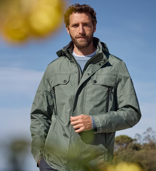 Ein Mann steht auf einer Frühlingswiese und trägt die Aquastop Reisejacke in Salbei.