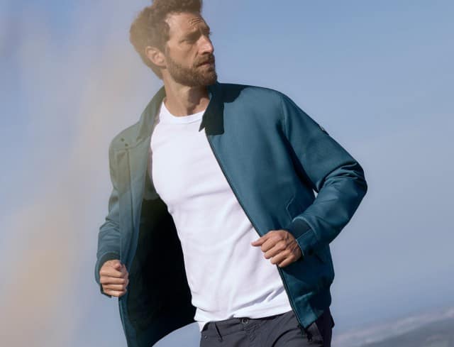 Ein Mann steht auf einem Feld und trägt einen Freizeitblouson in Rauchblau, darunter ein weißes Shirt.