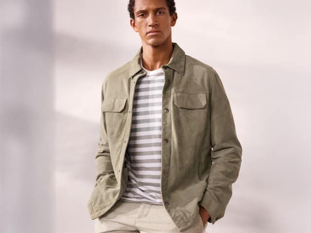 Ein Mann vor einem hellen Hintergrund trägt eine Velourslederhemdjacke in Salbei, darunter ein gestreiftes Leinenshirt.