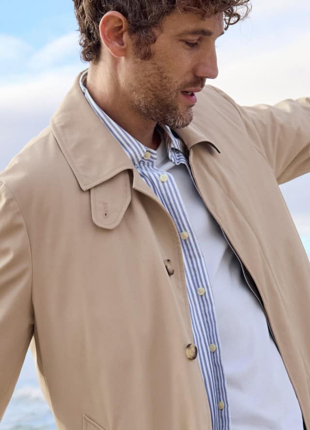 Eine Mann am Strand, trägt einen beigefarbenen Trenchcoat über einem hellen, gestreiften Hemd, was klassisch, leicht und entspannt wirkt.