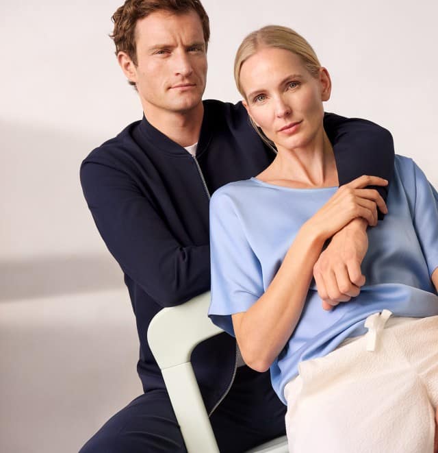 Ein Mann trägt einen Blouson in Marine mit passender Hose, eine Frau sitzt auf einem Stuhl und trägt ein hellblaues Shirt, dazu eine weiße Hose.