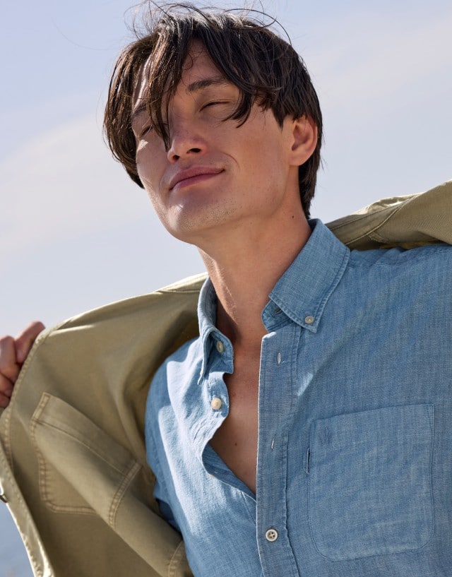 Ein Mann am Strand, trägt ein Hemd in Blau und darüber eine offene leichte Jacke in Khaki.