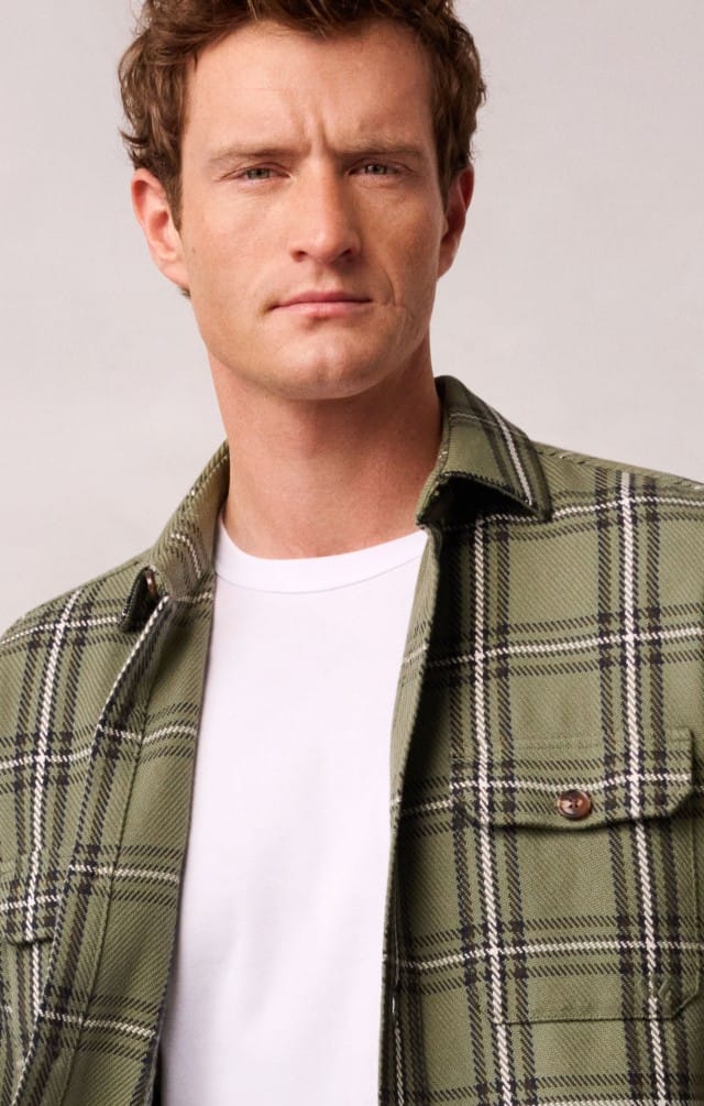 Ein Mann trägt ein offenes kariertes Hemd in Khaki, darunter ein weißes Shirt.