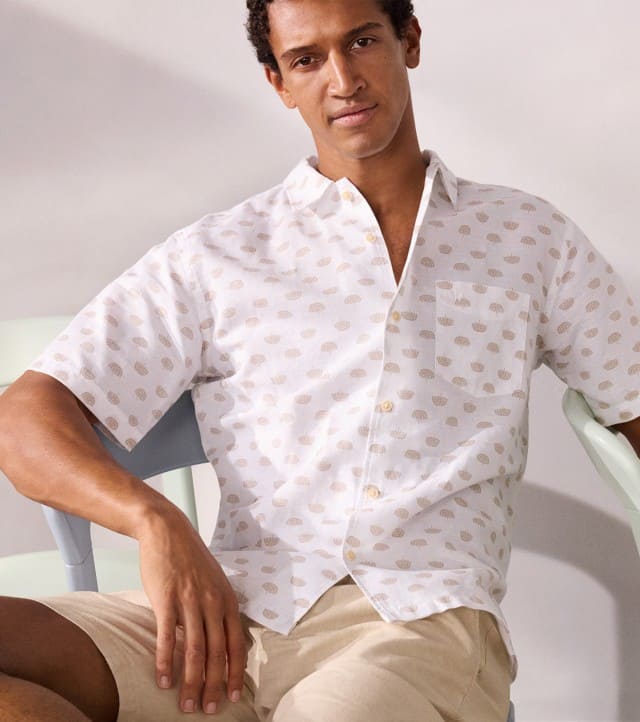 Ein Mann sitzt lässig auf eine Stuhl und trägt ein weißes Hemd mit Muster, dazu eine helle Hose.