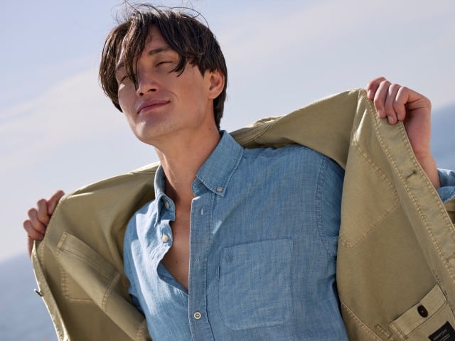 Ein Mann am Strand, trägt ein Hemd in Blau und darüber eine offene leichte Jacke in Khaki.