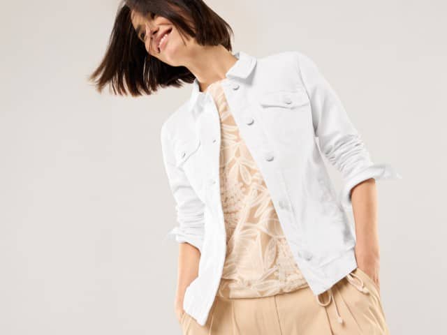 Eine Frau trägt eine weiße Jeansjacke, darunter ein Shirt und dazu eine Hose in Beige. 