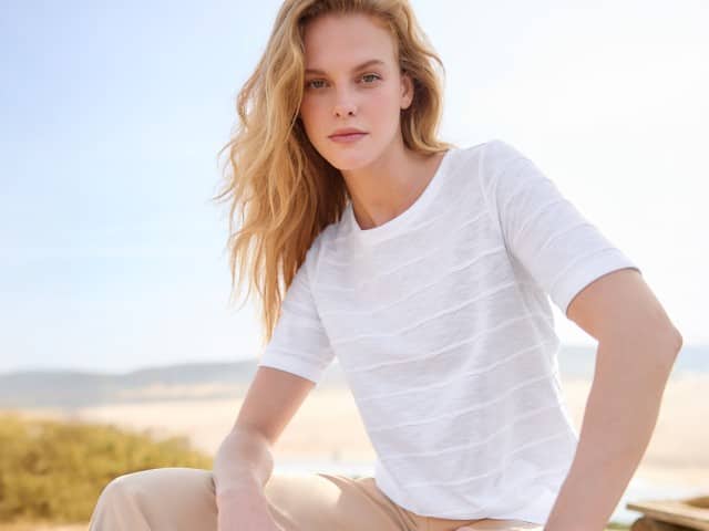 Eine Frau sitzt am Strand und trägt ein Baumwollshirt Streifenstruktur in Weiß.