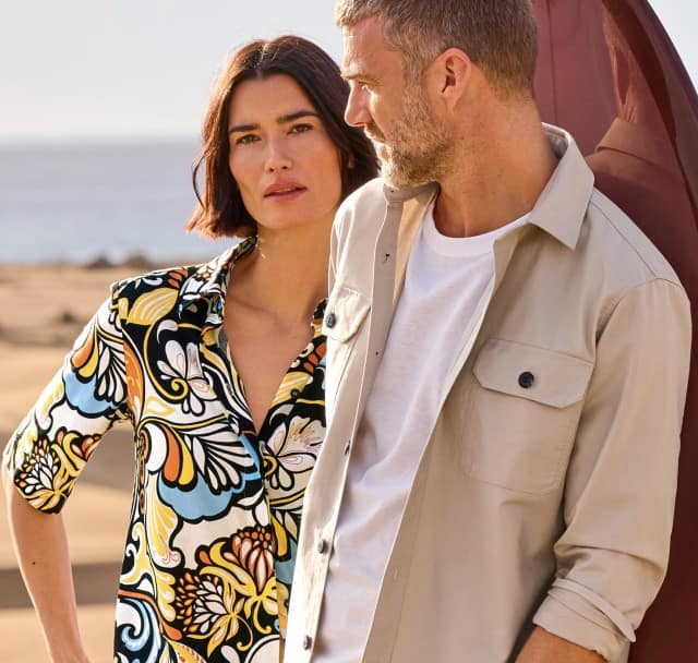 Eine Frau und ein Mann stehen in den Dünen. Sie trägt eine gemusterte Bluse und er eine helle Hemdjacke mit weißen Shirt darunter. 