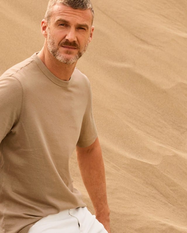 Ein Mann steht in den Dünen und trägt ein helles T-Shirt, dazu eine weiße Hose.