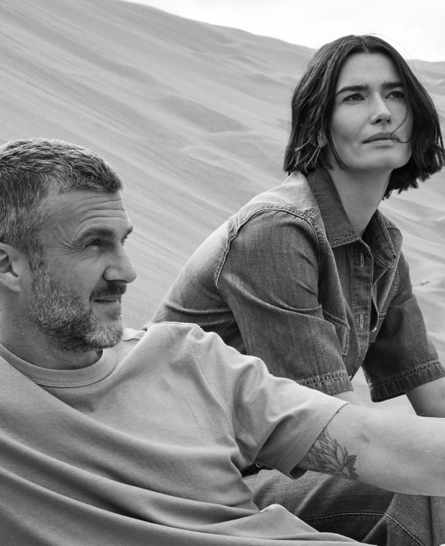 Zwei Personen sitzen in einer Sanddüne, einer trägt ein helles T-Shirt, die andere ein Jeanshemd.