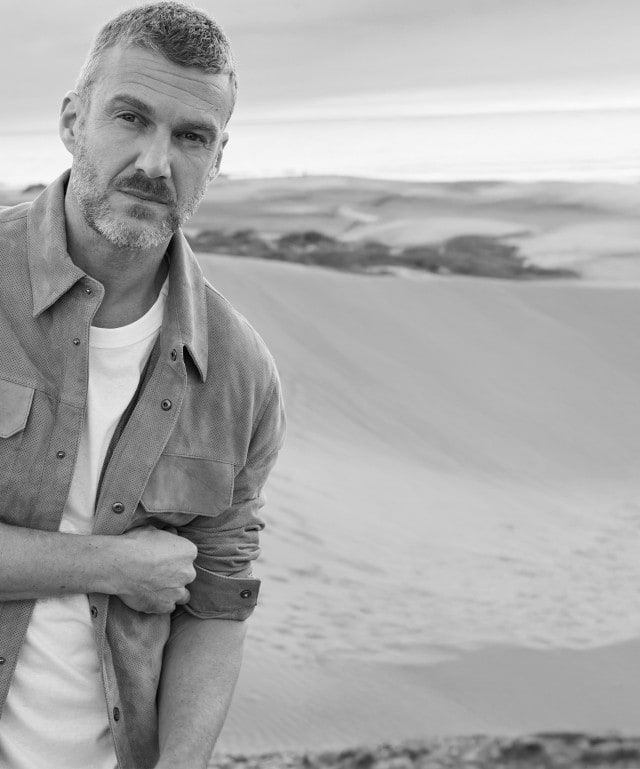 Ein Mann steht in einer Dünenlandschaft und trägt ein helles T-Shirt unter einer offenen Hemdjacke mit Brusttaschen.
