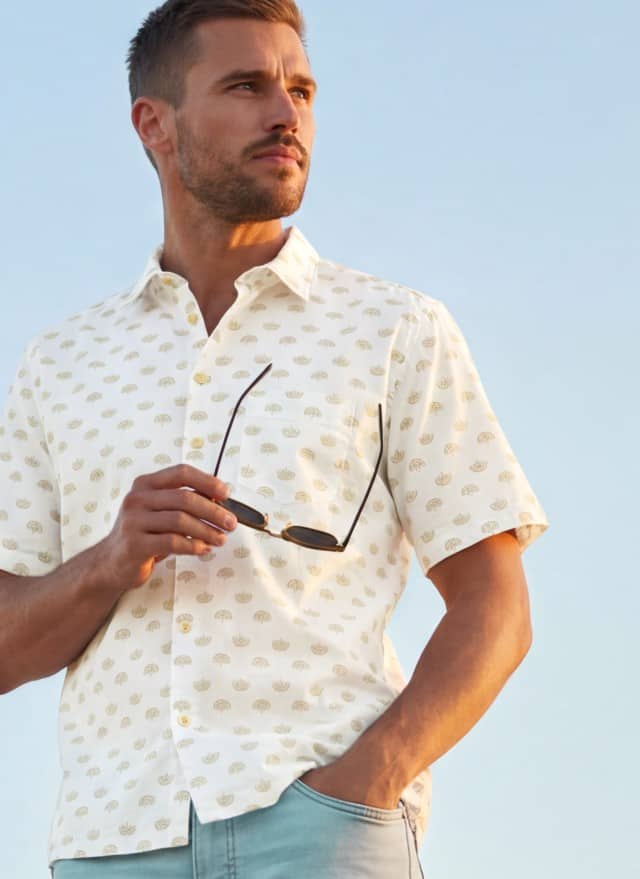 Ein Mann trägt ein weißgemustertes Kurzarmhemd und hält eine Sonnenbrille in der Hand.