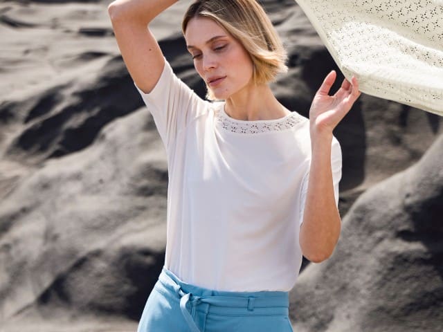 Eine Person trägt ein weißes Oberteil mit Lochstrickdetails am Ausschnitt und eine hellblaue Hose mit Bindegürtel – ein luftiger, sommerlicher Look.