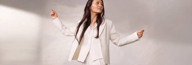 Frau trägt einen eleganten cremefarbenen Hosenanzug aus strukturiertem Stoff und posiert mit ausgebreiteten Armen in minimalistischer Umgebung. Der Stil wirkt modern und zeitlos.