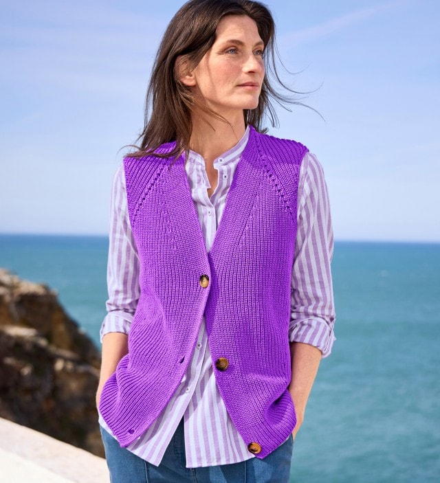 Eine Frau in einer gestreiften Bluse und Strickweste in Lila-Tönen steht lässig am Meer und schaut zur Seite.