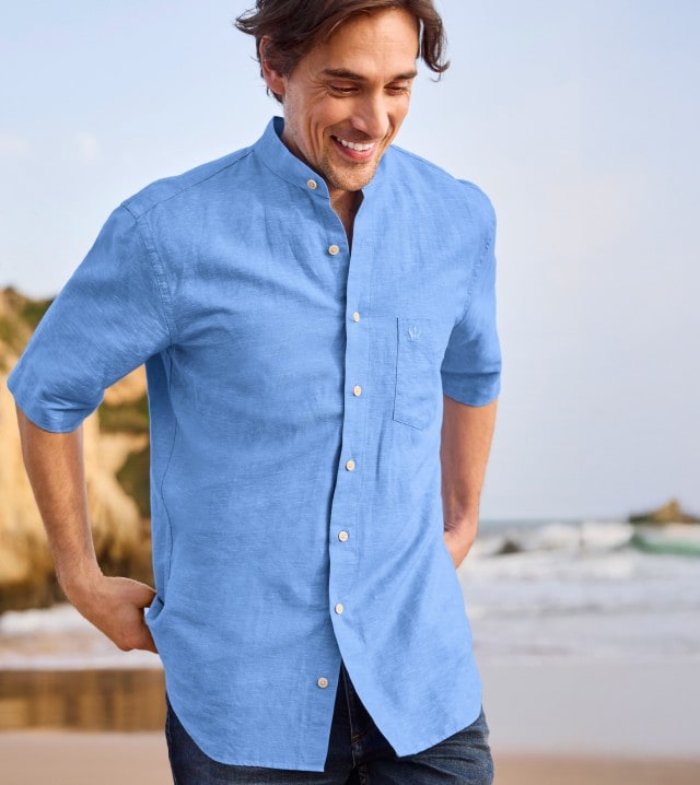 Ein Mann in einem blauen Kurzarm-Hemd steht am Strand und schaut lächelnd nach unten.