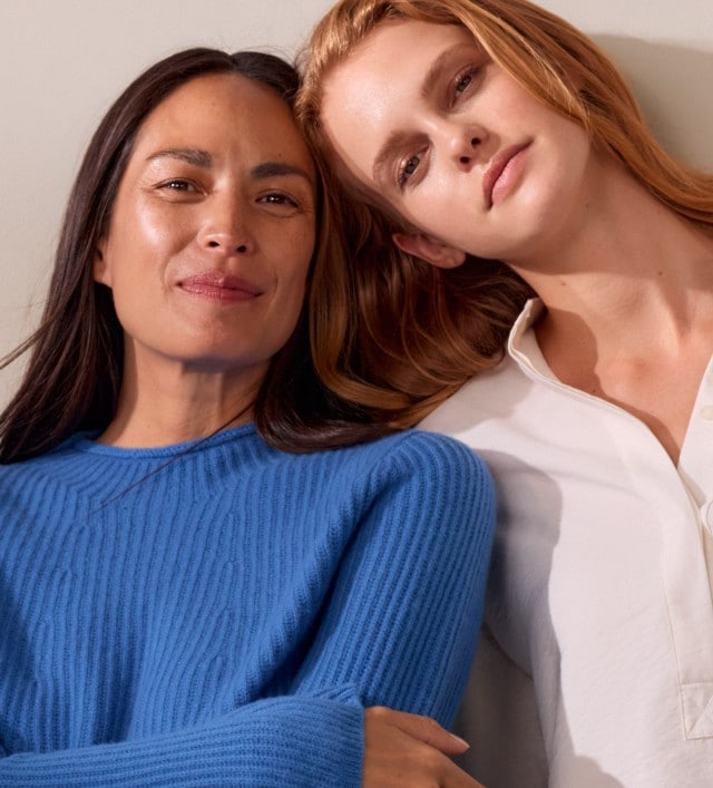 Zwei Frauen stehen nebeneinander, eine trägt einen blauen Strickpullover, die andere eine weiße Bluse mit Stehkragen und Knopfleiste.