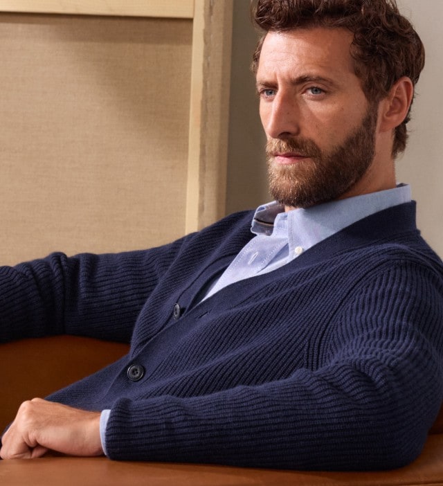 Mann trägt eine blaue Strickjacke über einem hellblau-gemusterten Hemd und grauer Stoffhose und sitzt locker in einem Sessel.