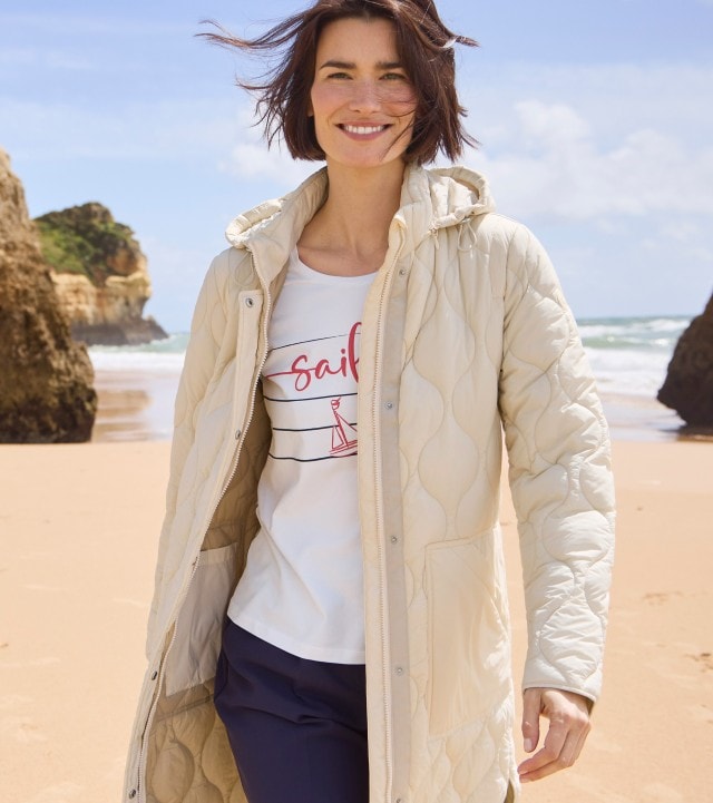 Frau in einer hellen Jacke und einem weißen bedruckten Shirt steht am Strand und schaut lächelnd in die Kamera.