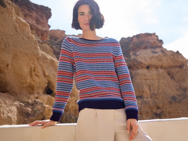 Frau in einem blau-rot-weiß gestreiften Strickpullover und heller Hose steht vor Felsen und schaut in die Kamera.
