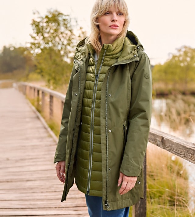 Eine Frau steht auf einem Steg und trägt einen Aquastop Parka 3-in-1 in der Farbe Oliv, dazu eine Jeans.