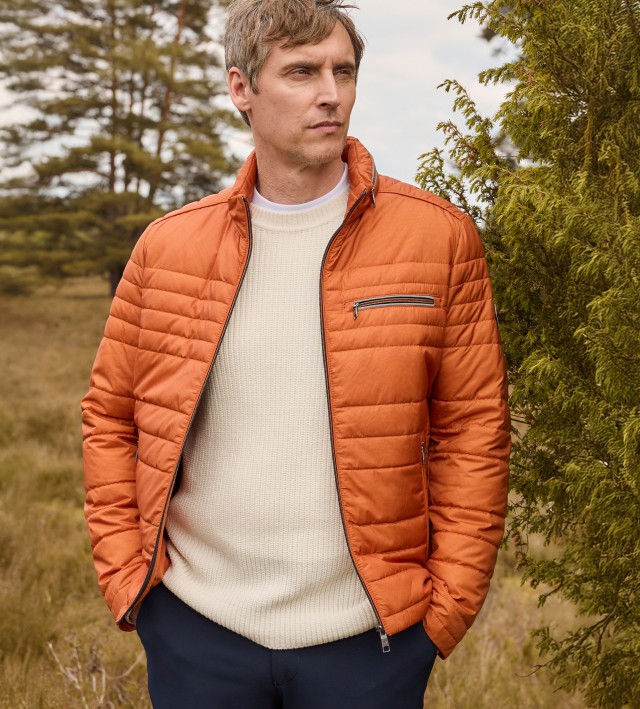 Ein Mann trägt eine Microfaser Leichtjacke in Orange, darunter einen hellen Strickpullover und dazu eine Hose in Marine.