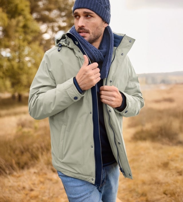 Ein Mann trägt  die Aquastop Wetterjacke in der Farbe Kitt, dazu einen blauen Schal und Mütze, sowie eine Jeans.