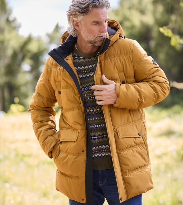 Ein Mann trägt eine senfgelbe Steppjacke über einem Strickpullover mit Norwegermuster und Jeans; der Stil wirkt warm, gemütlich und herbstlich.
