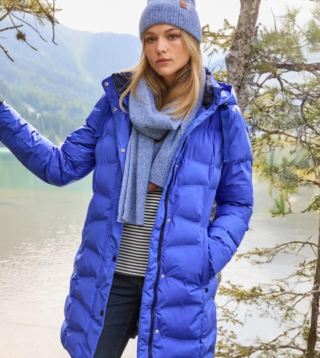 Eine Frau steht vor einem Bergsee und trägt den Aquastop Steppmantel 3D in Tintenblau, dazu ein gestreiftes Oberteil, eine dunkele Hose und eine passende Mütze und Schal. 