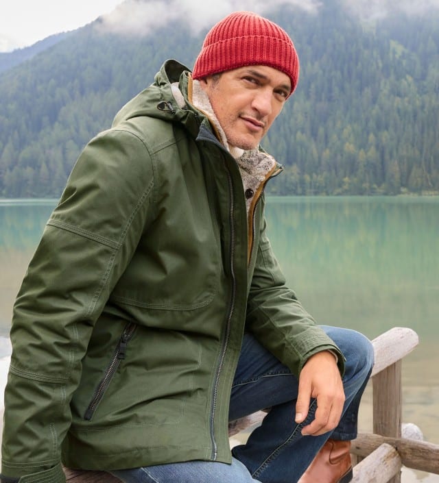 Ein Mann steht an einem Bergsee und trägt die Aquastop Winterjacke in Oliv, dazu eine Jeans und eine rote Mütze.