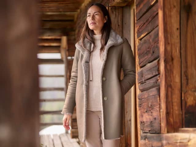 Eine Frau steht vor einer Hütte und trägt einen Rollkragen-Pullover, darüber den Premium Lammfell Wendemantel in Taupe.