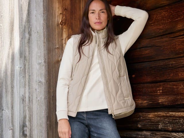 Eine Frau steht vor einer Holzwand und trägt die Wellensteppweste in Toffee/Creme, darunter ein weißes Sweatshirt und dazu eine Jeans.