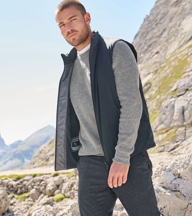 Ein Mann steht vor einem Gebirge und trägt einen grauen Strickpullover, darüber eine Weste in Marine und dazu eine dunkele Hose.