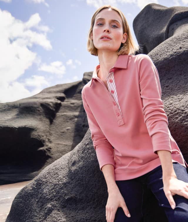 Eine Frau sitzt auf einem Felsen an der Küste und trägt einen Pullover mit Kragen in der Farbe Koralle.