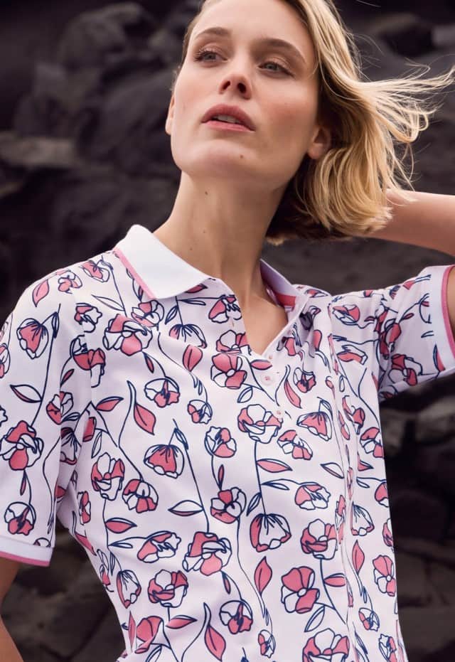 Frau mit blondem Haar in floral gemustertem Poloshirt vor einer dunklen Felslandschaft.
