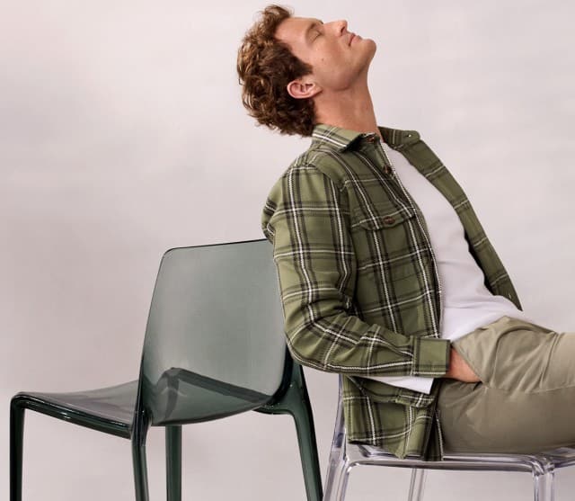Ein Mann sitzt entspannt auf einem Stuhl, trägt ein offenes kariertes Hemd in Khaki, darunter ein weißes Shirt und dazu eine passende Hose. 