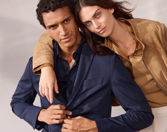 Ein Mann trägt ein Travel Anzug-Sakko in Marine, mit passendem Hemd dazu. Eine Frau in einem Ziegenvelours Perfo Blouson in Caramel, mit einer Polo Bluse aus Seide in der Farbe Praline darunter.