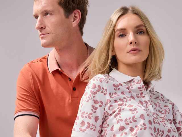 Ein Mann trägt ein Poloshirt in Orange, rechts daneben eine Frau in einem Poloshirt mit Muster Tulpenspiel in der Farbe Rosenholz.