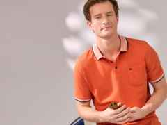 Mann in orangefarbenem Poloshirt lehnt an einer weißen Wand, hält eine Sonnenbrille in der Hand, schaut leicht lächelnd zur Kamera.