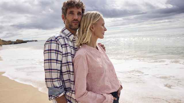 Ein Mann und eine Frau stehen am Strand, blicken aufs Meer. Der Himmel ist wolkig. Beide tragen legere Kleidung.